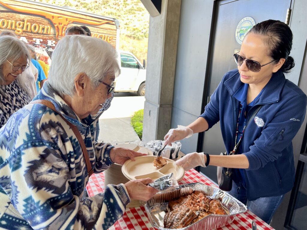 A woman in a blue shirt and plastic gloves is holding a small piece of pink salmon on a plastic fork. She is serving it to another woman with gray hair and a blue and white fleece shirt. That woman is holding a white paper plate. A silver aluminum pan of salmon sits on top of a red and white checkered table cloth at the bottom of the picture.