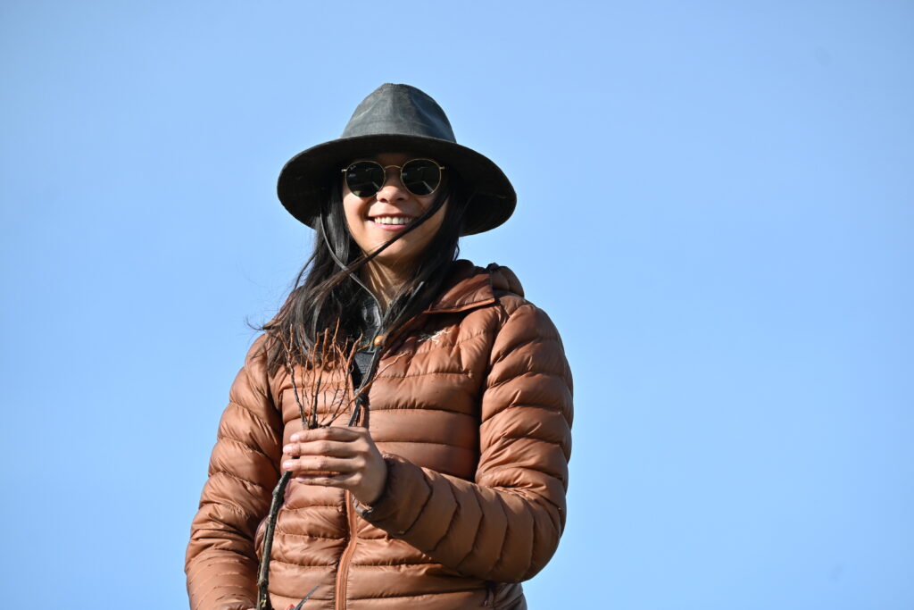 A woman with black sunglasses, a grey hat and brown jacket holds a single brown grafted grapevine against a blue sky. 