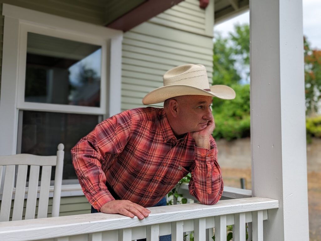 Curt Westberg stands on his white porch with a red plaid shirt and white cowboy hat on.