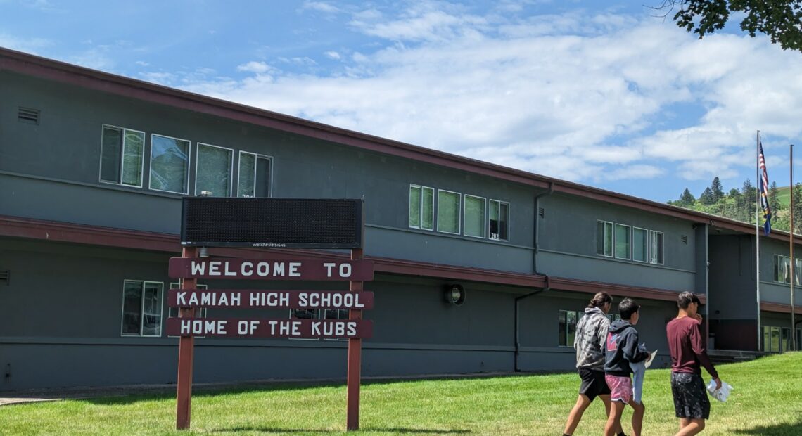 Three students in shorts walk near a maroon sign on a green lawn that reads "Welcome to Kamiah High School, home of the Kubs."