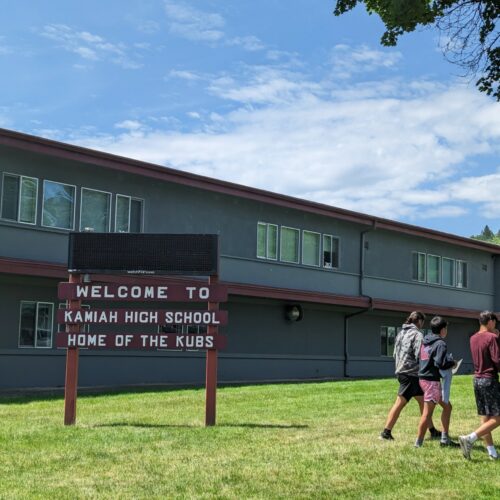 Three students in shorts walk near a maroon sign on a green lawn that reads "Welcome to Kamiah High School, home of the Kubs."
