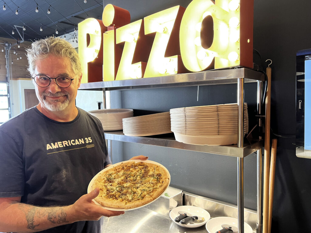  A man in a blue shirt that says "American 35" on it stands holding a pizza with brown fig sauce on it. He is standing in front of a yellow sign that says "Pizza" and a silver shelf with three piles of white plates and three pizza cutting tools on it.
