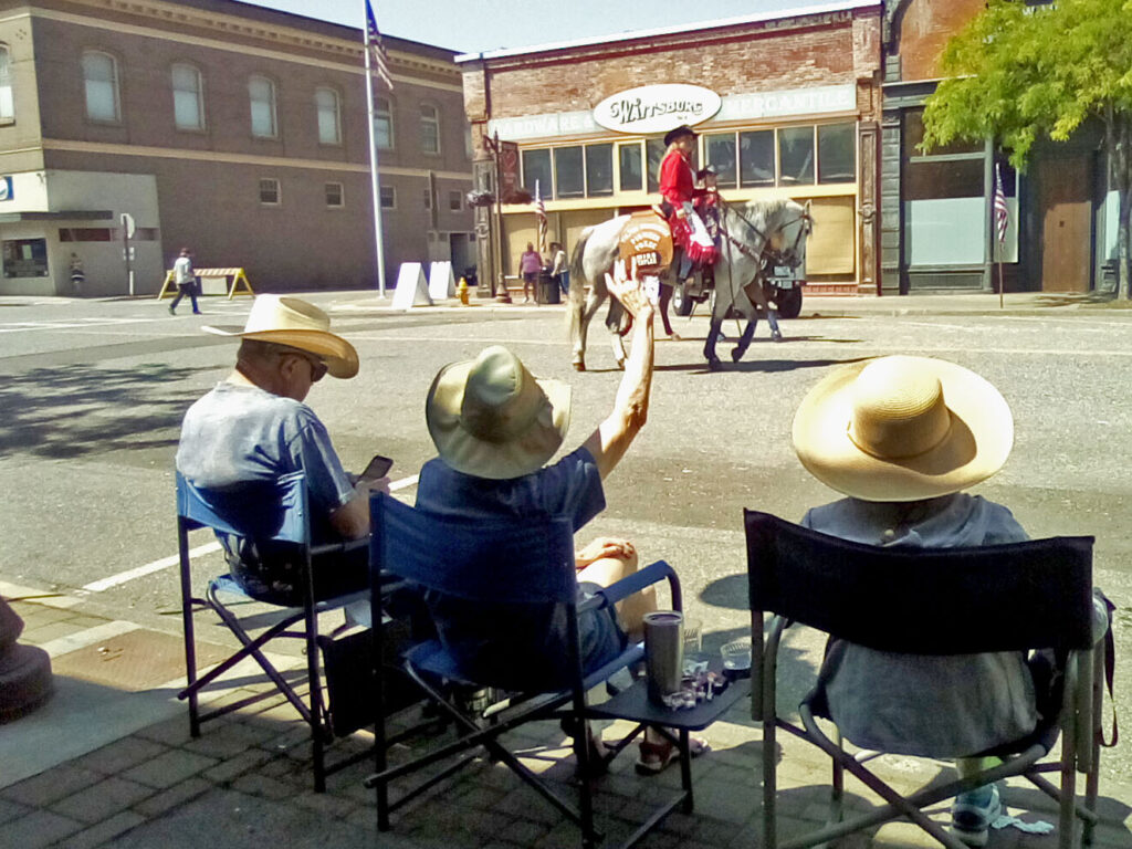 Three people in tan cowboy hats sit in blue folding chairs. They are wearing grey T-shirts. They are sitting on a sidewalk. In the middle of the street, a woman in a red cowboy outfit is riding a grey horse. A building behind her has a white and black sign that says, "Waitsburg."