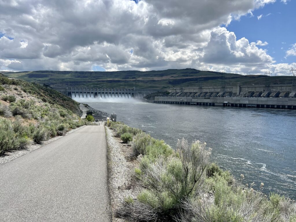 A grey sidewalk is on the left side of the photo. It's surrounded by green shrubs. To the right of the shrubs is a blue river. In the background, white water spills over a cement dam. The sky has lots of grey clouds.