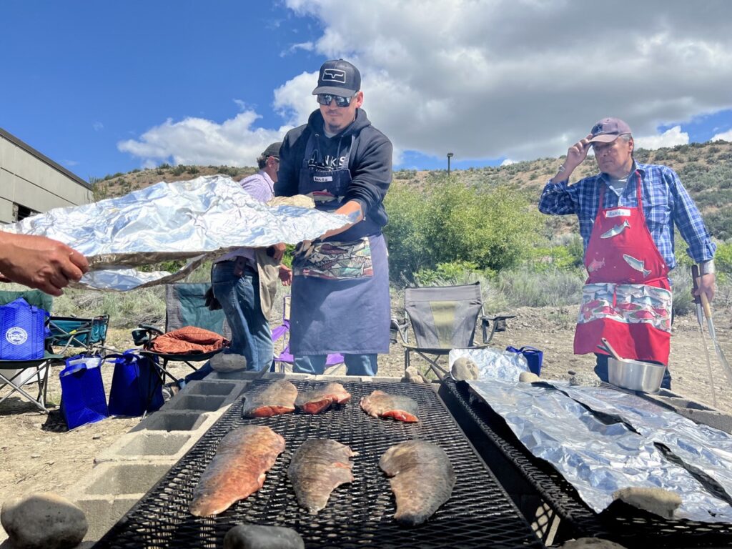 A man in a black baseball cap, sunglasses, a black hoodie and a blue apron holds a large piece of silver foil over six pink and grey salmon filets. The filets are on a black grill that's on top of cement blocks to form a fire pit. In the background, a man in a blue plaid shirt, red apron and blue baseball cap watches. There are clouds in the sky.