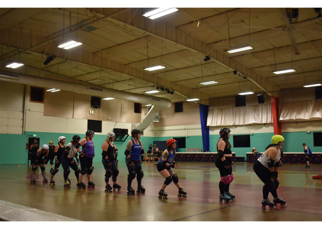 Roller derby skaters move in a line on a gym floor beneath a high ceiling with fluorescent lights. They wear helmets, knee and elbow pads and roller skates. Some crouch, while others stand upright.