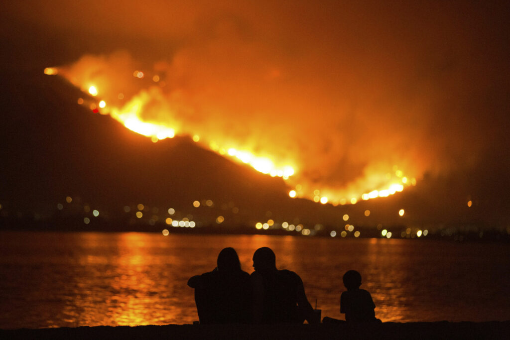 A silhouette of a family sitting on a lake watching a fire burn across the horizon.