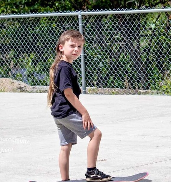 A little boy with long light brown hair, a black shirt and grey shorts stands on a black skateboard. Behind him is a chain link fence.