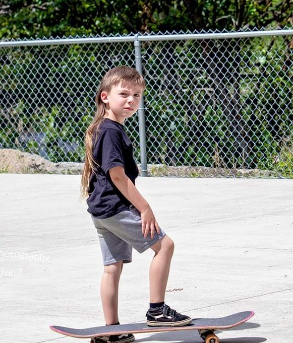 A little boy with long light brown hair, a black shirt and grey shorts stands on a black skateboard. Behind him is a chain link fence.