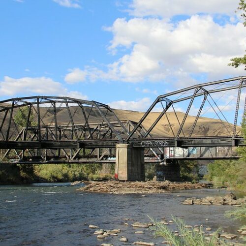 in the bottom third of the photo, there is a blue river with tan rocks in it. At grey railroad bridge is crossing the river. On the right hand side of the photograph, there are green bushes and trees. In the background, there are tan hills, a blue sky and white clouds at the top of the photo.