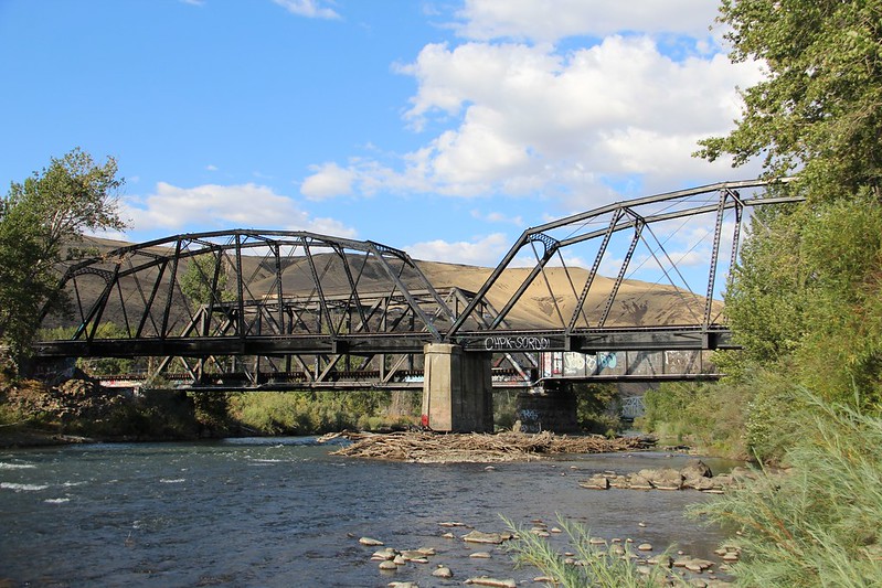 in the bottom third of the photo, there is a blue river with tan rocks in it. At grey railroad bridge is crossing the river. On the right hand side of the photograph, there are green bushes and trees. In the background, there are tan hills, a blue sky and white clouds at the top of the photo.