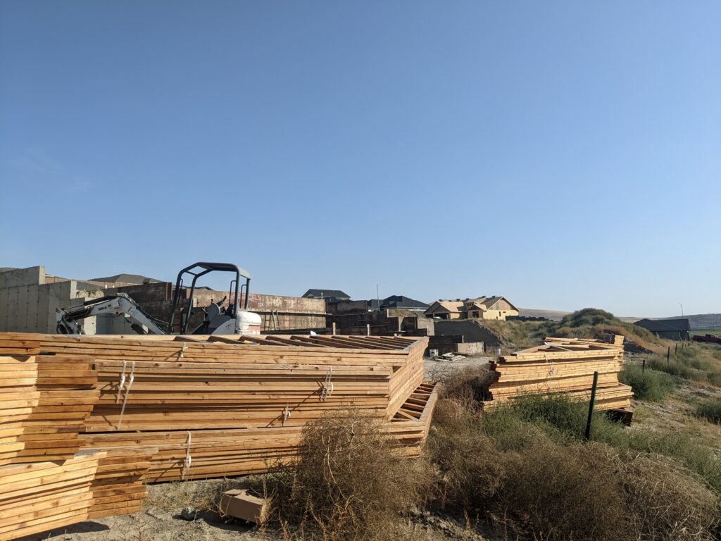 Piles of lumber are framed by a blue sky with houses being built in the distance.