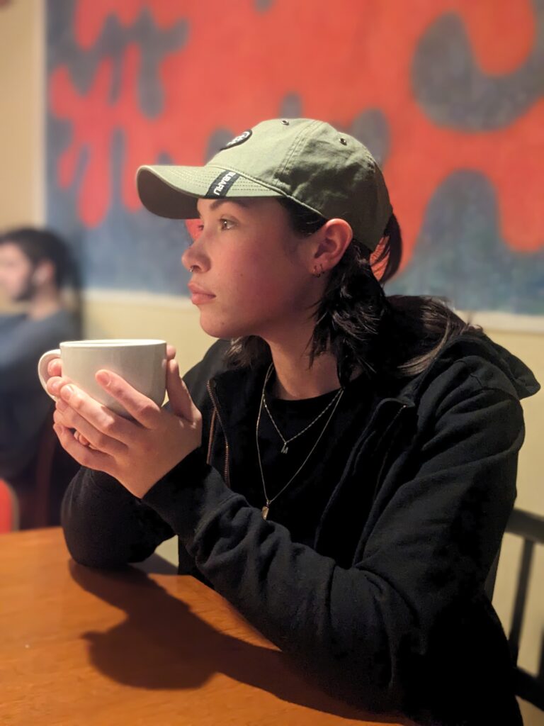 A woman in a black jacket and green ball cap sits at a table holding a white mug filled with tea. 
