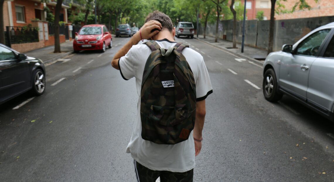 A young man with a black backpack walks down a neighborhood street.