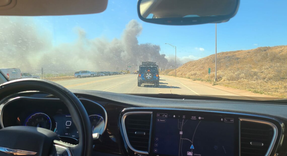 The picture is taken from the inside of a black car. In the front of the picture there is a steering wheel and navigation guid on the car. In front of the car, there is a blue jeep stopped in traffic. In front of the jeep, there is a big plume of smoke going across the road. On the left hand side of the picture, there is a small grassy hill.