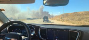 The picture is taken from the inside of a black car. In the front of the picture there is a steering wheel and navigation guid on the car. In front of the car, there is a blue jeep stopped in traffic. In front of the jeep, there is a big plume of smoke going across the road. On the left hand side of the picture, there is a small grassy hill.