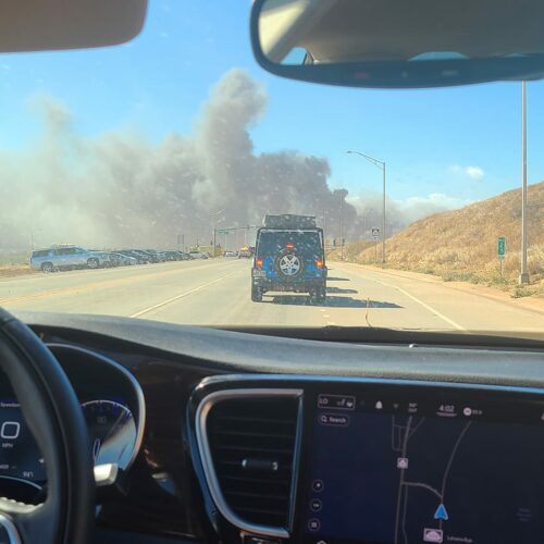 The picture is taken from the inside of a black car. In the front of the picture there is a steering wheel and navigation guid on the car. In front of the car, there is a blue jeep stopped in traffic. In front of the jeep, there is a big plume of smoke going across the road. On the left hand side of the picture, there is a small grassy hill.