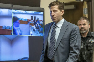 Bryan Kohberger wears a grey suit and striped grey tie as an officer in uniform walks behind him into the courtroom.