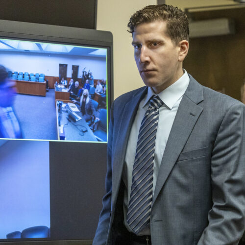 Bryan Kohberger wears a grey suit and striped grey tie as an officer in uniform walks behind him into the courtroom.