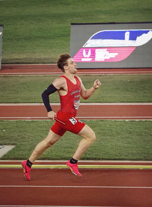 A man in a red tank top, red shorts and red shoes is running on a brown track. There is green astroturf and brown turf on the ground behind him.