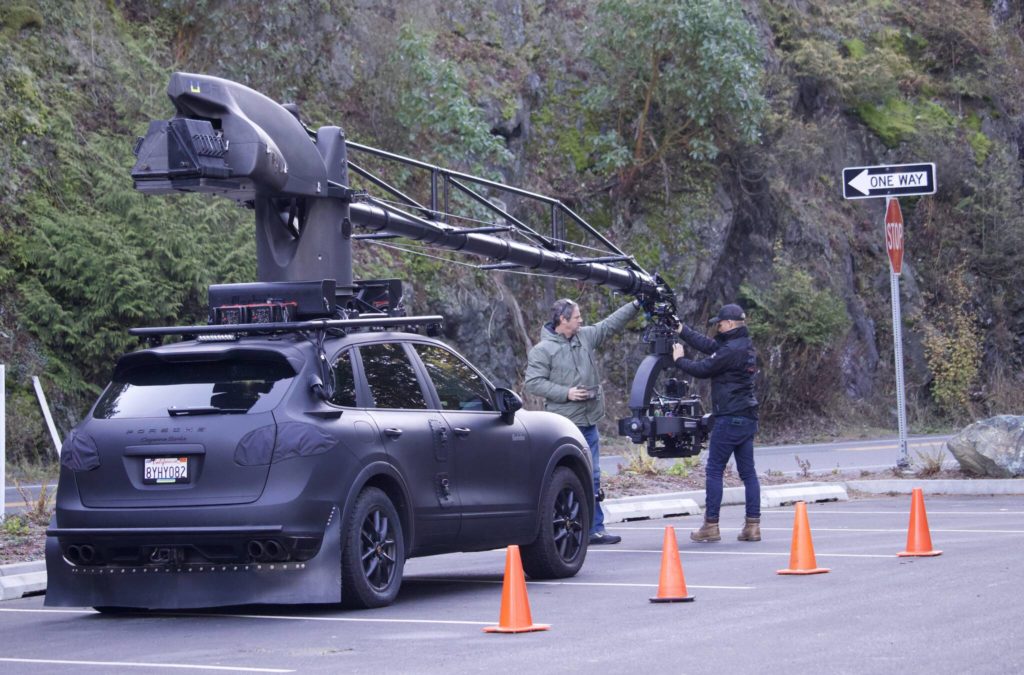 Orange cones sit on the road beside a black Porsche with a black crane camera.