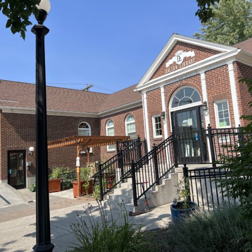 A red brick building with black stair railings. A small brown pergola is in front of the library. A few green plants are underneath the pergola. An evergreen tree is at the side of the red brick building. A black lamppost is in front of the building.