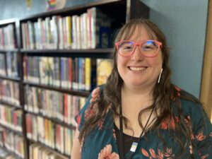 A woman with pink glasses and long brown hair stands in front of a library bookshelf.