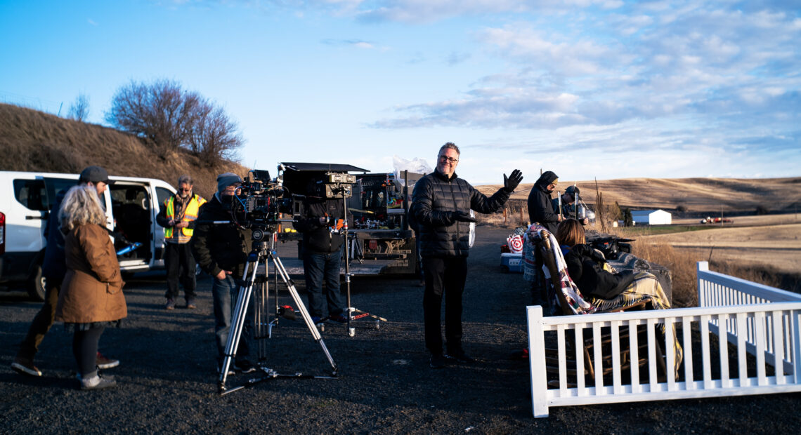 A camera is set up behind a white picket fence while crew members dressed in black fill the scene against a wheat field and blue sky.