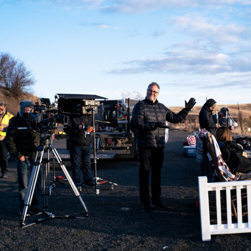 A camera is set up behind a white picket fence while crew members dressed in black fill the scene against a wheat field and blue sky.