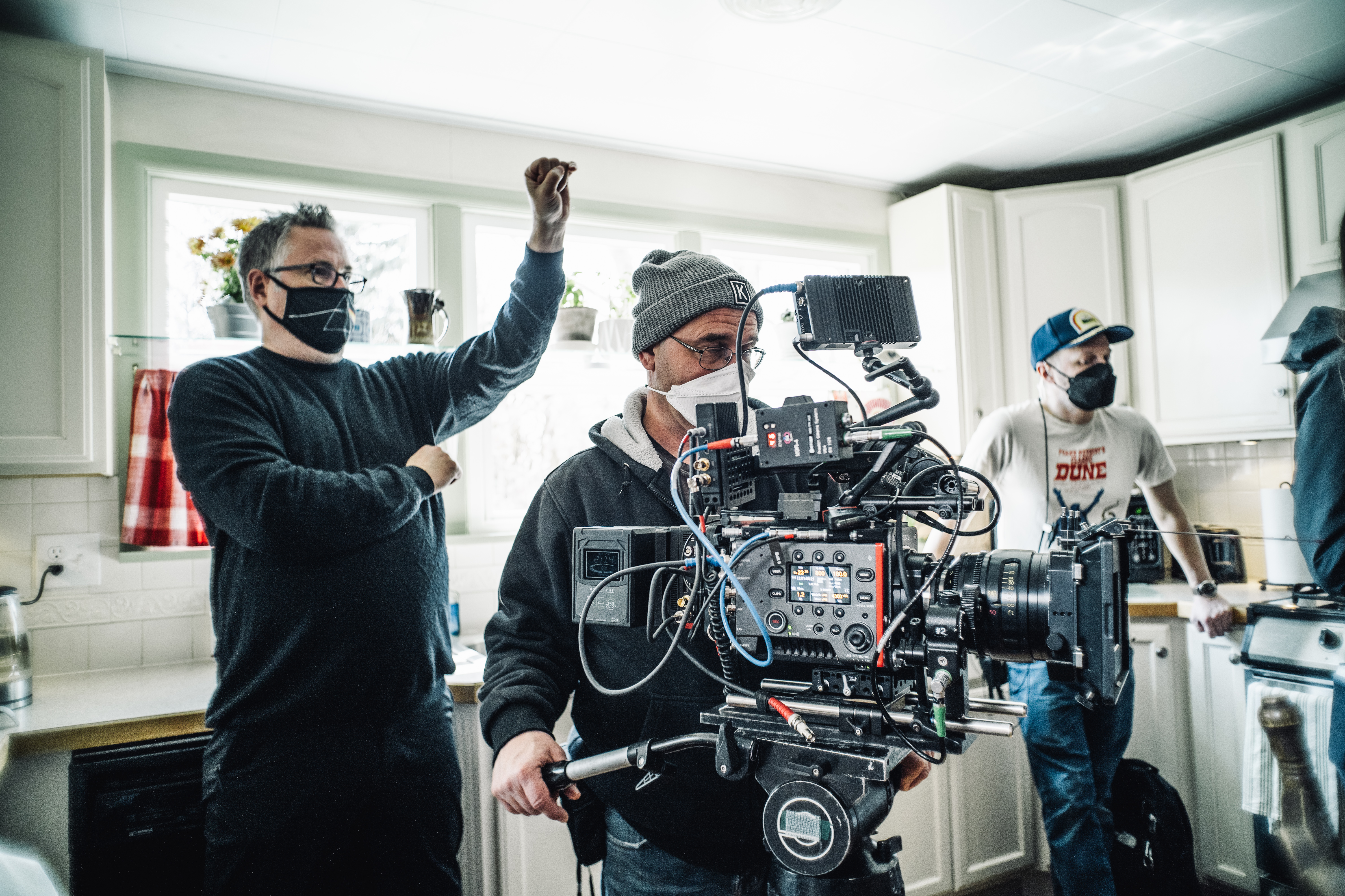 A large camera sits in a kitchen with cast and crew behind. The director is giving a hand signal.