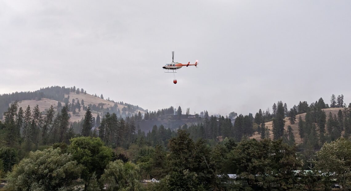 A helicopter with a bucket of water flies above a tree covered hillside with smoke billowing in the distance.