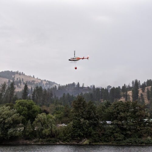 A helicopter with a bucket of water flies above a tree covered hillside with smoke billowing in the distance.