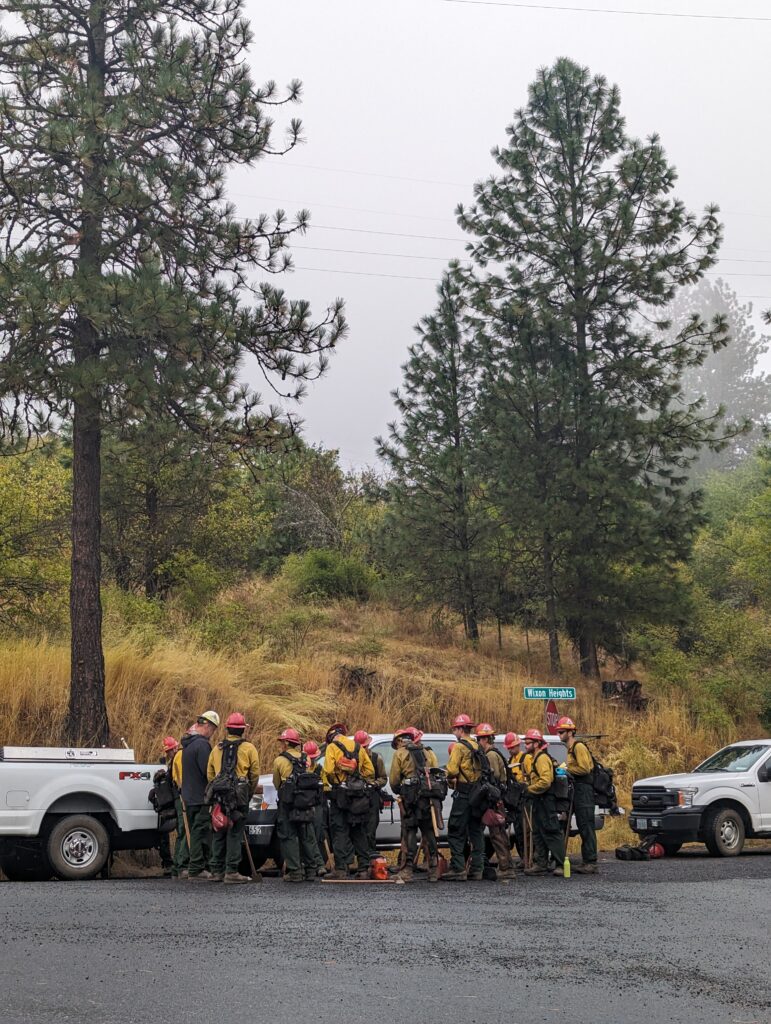 Firefighters in full gear and yellow helmets stand in a circle near two white trucks against two large evergreen trees. 