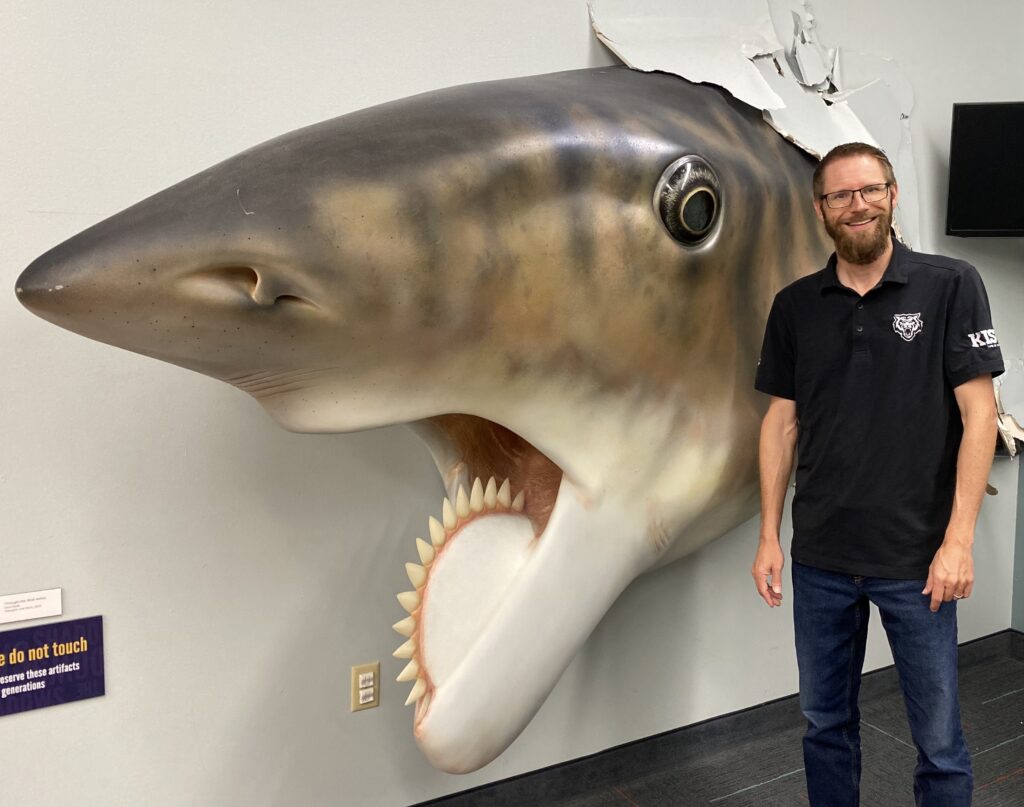 Portrait of Dr. Leif Tapanila. A huge helicoprion shark model is bursting through the wall behind him.