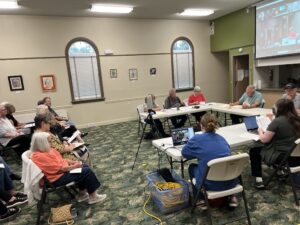 People sit around white tables that are forming a horseshoe. There is a projector in the middle with people on zoom on a screen behind the table. Other people sit in chairs, holding notebooks and paper.