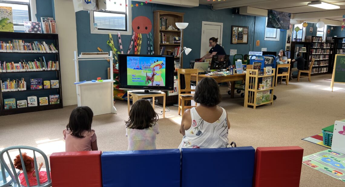 Two little girls in pink and white shirts sit on plush red and blue kid-sized chairs with their caretaker, who is in a white shirt. They are in a library with windows, books and a TV with a cartoon character on it.