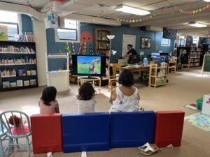 Two little girls in pink and white shirts sit on plush red and blue kid-sized chairs with their caretaker, who is in a white shirt. They are in a library with windows, books and a TV with a cartoon character on it.