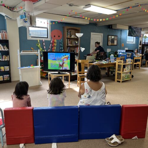 Two little girls in pink and white shirts sit on plush red and blue kid-sized chairs with their caretaker, who is in a white shirt. They are in a library with windows, books and a TV with a cartoon character on it.