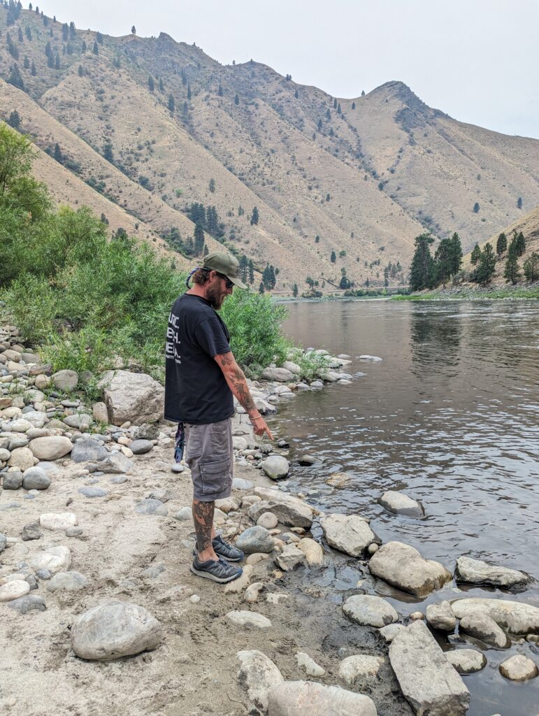 A man with a green hat, a beard, black shirt and cargo shorts points down at rocks on the shore of the Salmon River in Riggins, Idaho.