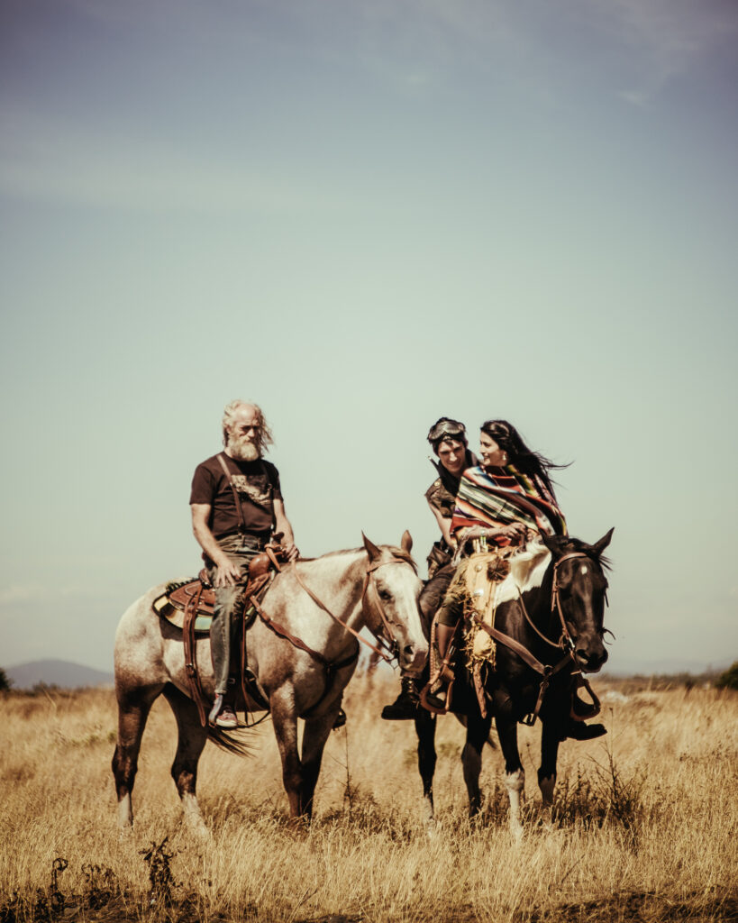 A man and a woman dressed in traditional clothing sit on a black horse next to a man with grey hair on a brown horse in a golden field. 