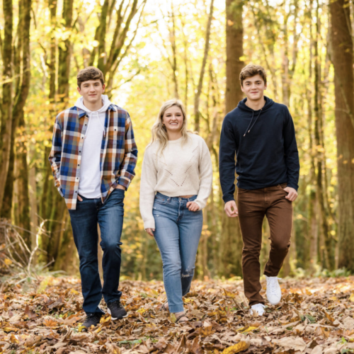 Two young men and a young woman in fall clothes stroll toward the camera between autumn trees.