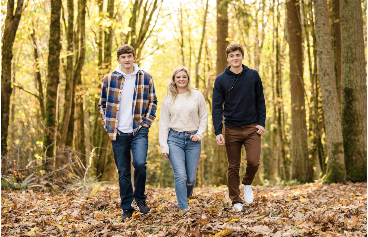 Two young men and a young woman in fall clothes stroll toward the camera between autumn trees.