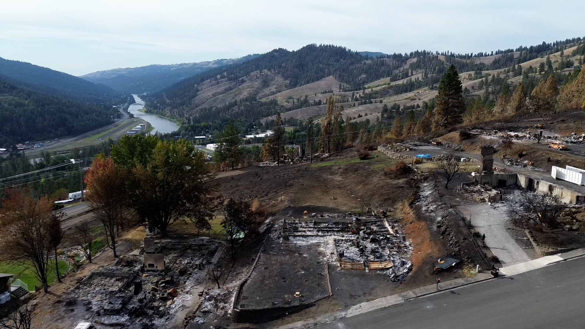 A drone photo shows a bird's eye view of the Wixson Heights neighborhood where several homes have been reduced to a sooty rubble. Tree covered mountains and a river are seen in the distance.