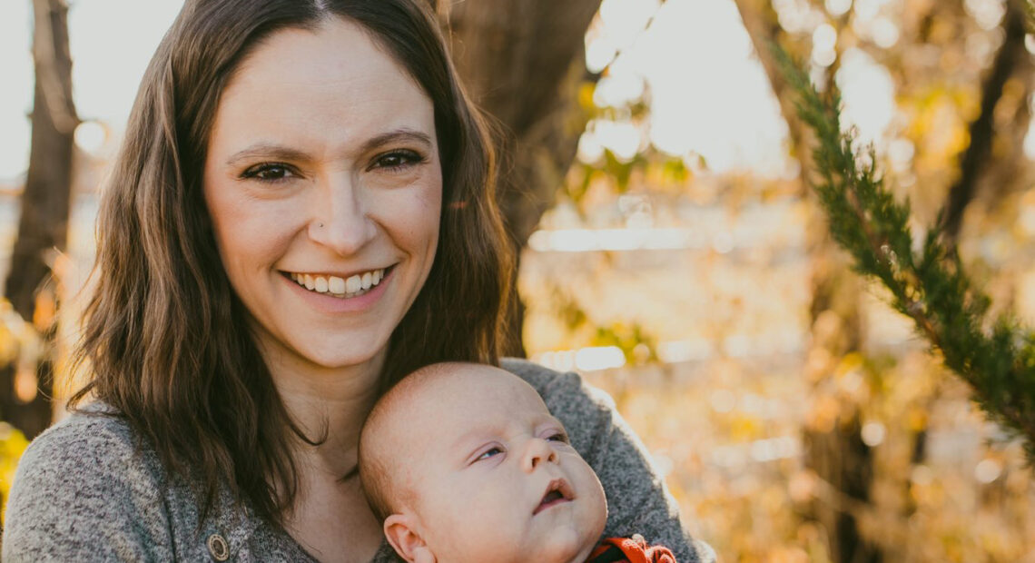 A woman and her baby are pictured in front a blurred forest scene near sunset. She's smiling toward the camera. She has shoulder-length brown hair that's parted in the center and is wearing a grey sweater. Her baby looks toward the right and has chubby cheeks and is wearing a red and black plaid long-sleeve top and blue jeans.