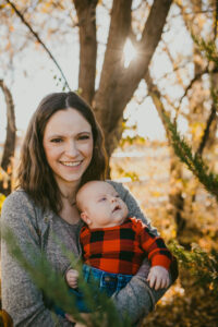 A woman and her baby are pictured in front a blurred forest scene near sunset. She's smiling toward the camera. She has shoulder-length brown hair that's parted in the center and is wearing a grey sweater. Her baby looks toward the right and has chubby cheeks and is wearing a red and black plaid long-sleeve top and blue jeans.