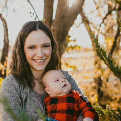 A woman and her baby are pictured in front a blurred forest scene near sunset. She's smiling toward the camera. She has shoulder-length brown hair that's parted in the center and is wearing a grey sweater. Her baby looks toward the right and has chubby cheeks and is wearing a red and black plaid long-sleeve top and blue jeans.