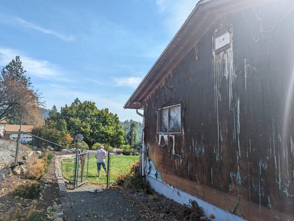 A home with a burned wall sits beside a stone path under a blue sky with green grass nearby. 