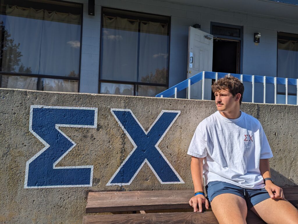 A young man with dark hair and a mustache sits on a bench with the Sigma Chi letters in bright blue painted nearby.