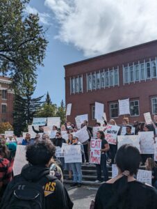 Students hold protest signs in front of a brick building flanked by large trees. One of the signs reads "eat the admins."