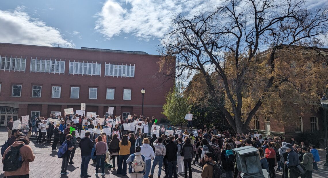 A crowd of students gather as students facing the crowd hold signs at a student union protest in front of a brick building under a blue sky.